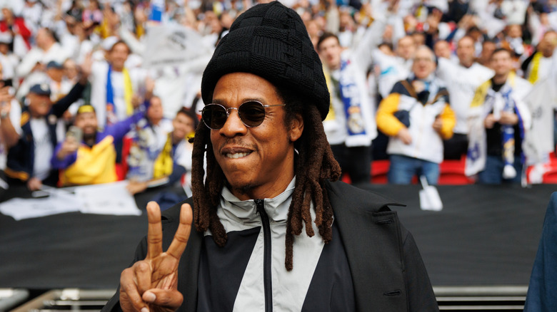 Jay-Z holding up a peace sign on a soccer field in front of cheering fans.