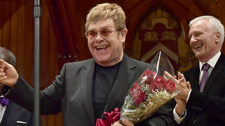 A smiling Elton John holding a boquet of roses onstage.