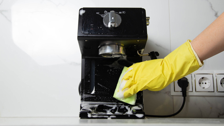 A girl s hand in a yellow glove washes a black coffee machine with a sponge with foam and cleaning agent in the kitchen against a background of a white wall with porcelain tiles.