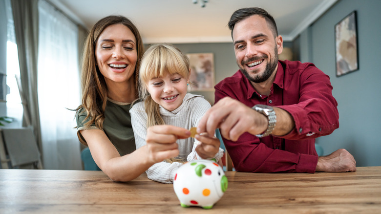 Young parents and daughter inserting coins in a piggybank at home.