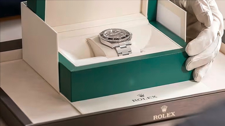 a white-gloved left hand holds open a turquoise box containing a Rolex watch