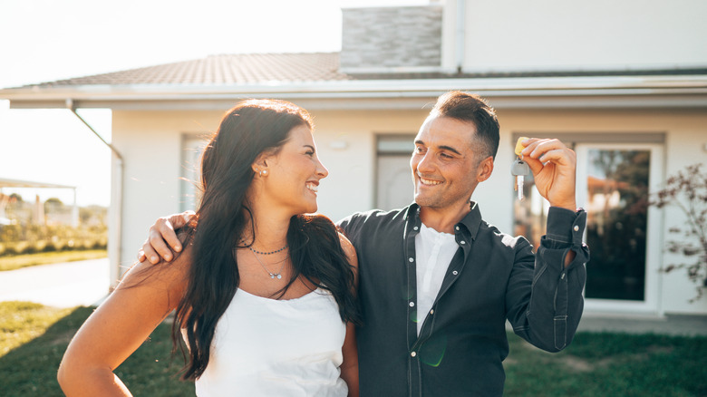 A happy couple smiling holding keys up keys in front of a sunlit home.