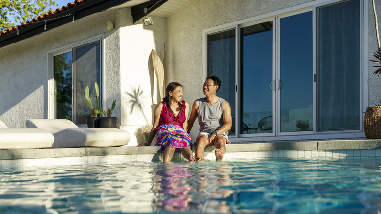 A smiling couple sitting poolside in their backyard with their feet in the water.