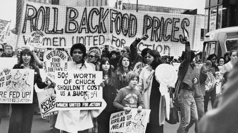 A 1970s crowd protests high food prices in city street, holding handmade signs