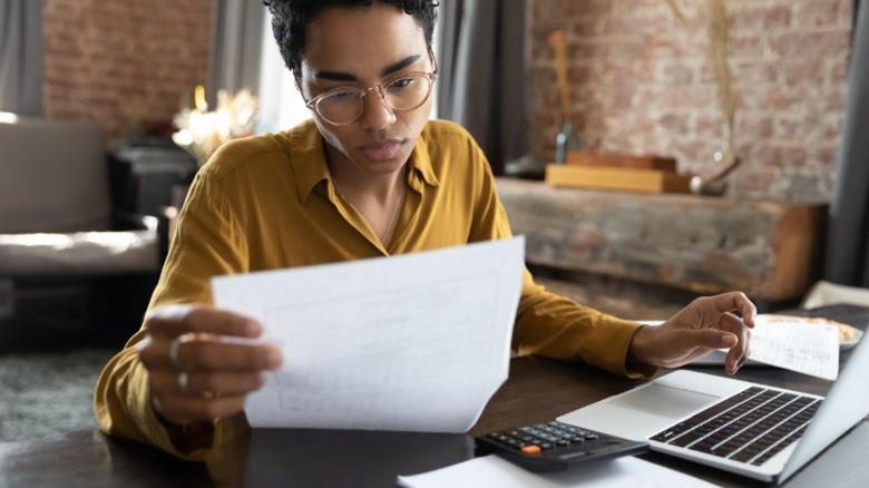 A young person looking at a sheet of paper while sitting at a desk in front of a laptop