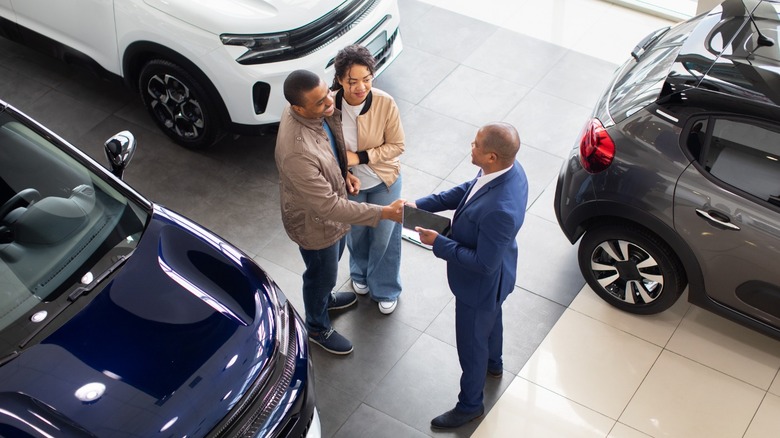 A young couple shopping for cars at a dealership and shaking hands with a car salesman.