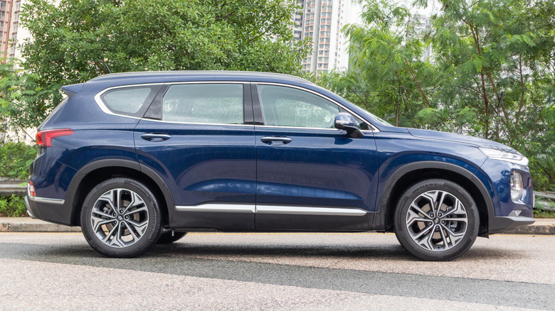 A side view of a dark Hyundai Santa Fe parked outdoors near trees with buildings in the background.