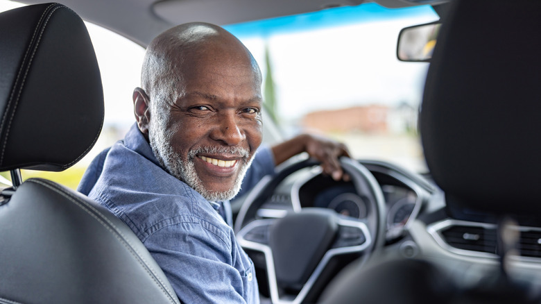A smiling senior man sitting in the driver's sear of a car looking toward the camera in the back seat.