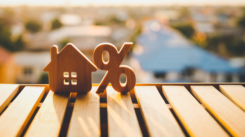 Wooden figurines of a house and percentage sign on a deck with the sun setting over a neighborhood behind them