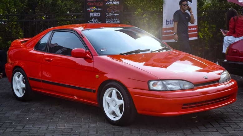 Red Toyota Paseo coupe with white wheels parked at an outdoor car meet