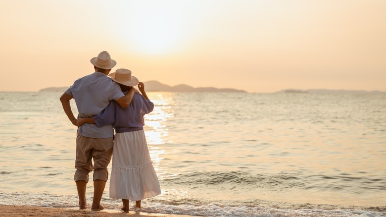 A retired couple standing on a beach watching the sunset.