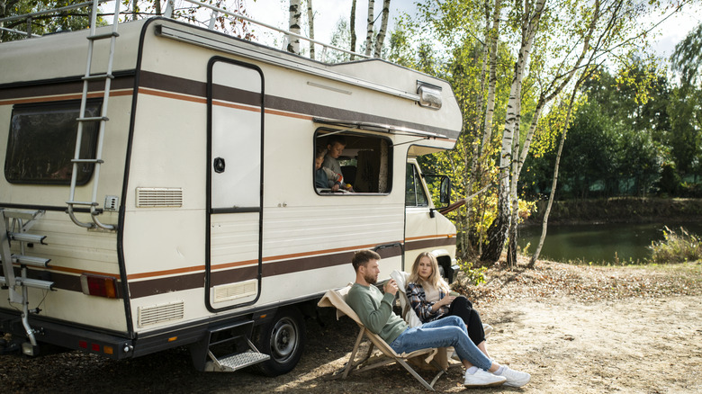 A couple sitting in front of an RV with their kids looking outside from the RV window.