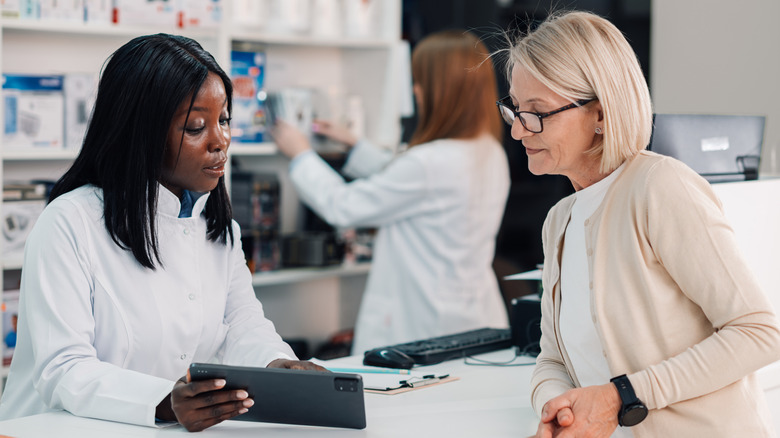 A pharmacist behind a counter at a pharmacy showing a retiree something on a tablet.