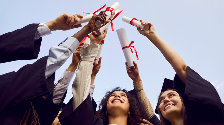 Happy graduate students standing in a circle in black robes and raising their diplomas up to the sky outdoor at graduation ceremony.