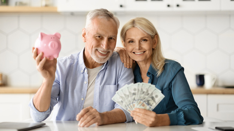 A smiling retired couple sitting together with the man holding up a small pink piggy bank and the woman fanning out paper money.