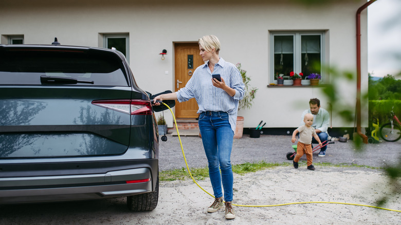 A woman charging electric car in front of house, holding smartphone.