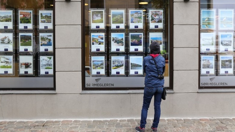 A person standing on a sidewalk looking at real estate listings posted in a business window.