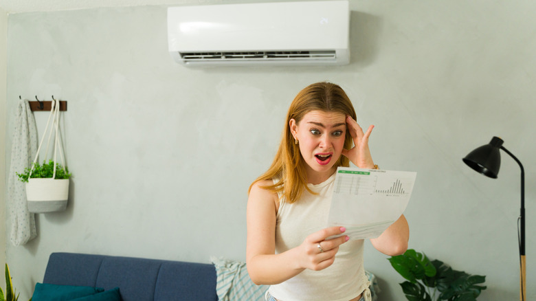 Stressed woman looking at expensive electricity bill while standing in living room with air conditioning unit