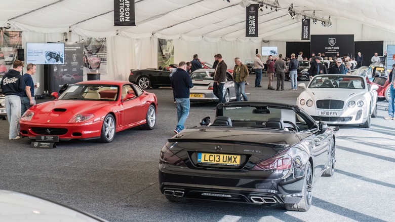 Luxury and sports convertibles displayed at an indoor car auction, with attendees walking among vehicles including a Ferrari and a Bentley