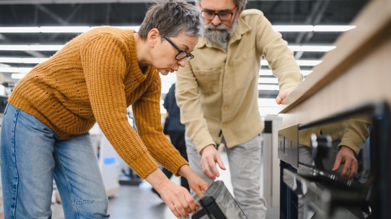 An older couple checking out a new oven in an appliance store by opening the door and looking inside.