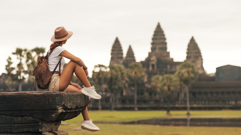 Solo female traveler sitting on ancient stone, admiring Angkor Wat temple in Cambodia.