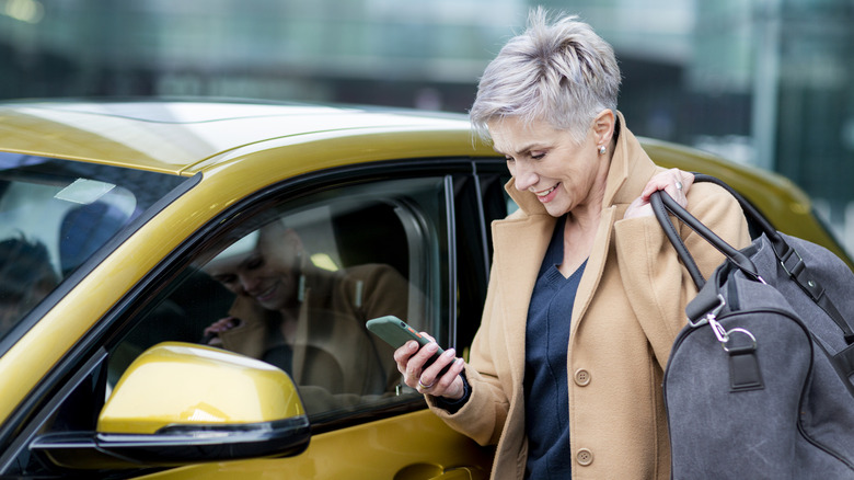 Woman holding luggage next to car while checking phone