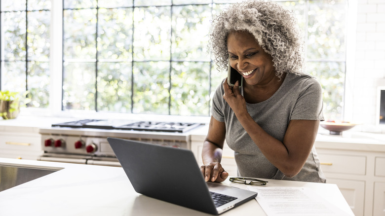 Senior woman using phone and computer in kitchen