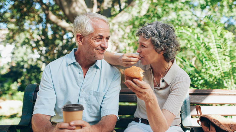 A smiling senior couple sitting with coffee and a muffin on a park bench on a sunny day