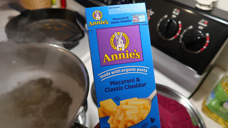 An open box of Annie's mac and cheese with a boiling pot of water on the stove in the background.