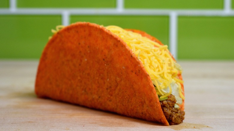 Crunchy Taco from Taco Bell filled with seasoned ground beef, shredded lettuce, and shredded cheese inside a bright orange hard shell, placed on a wooden surface with a green-tiled background