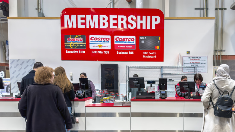 A Costco membership counter where customers are being assisted by employees, with a large sign displaying membership options.