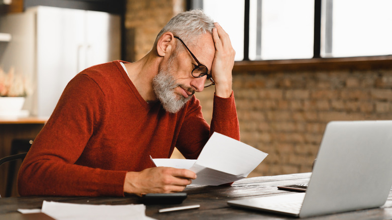 A Gen X man reading a bill with a concerned expression in front of an open laptop.