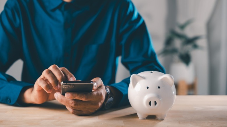 A man in a blue button-down shirt using his cell phone while sitting at a table with a piggy bank on it.