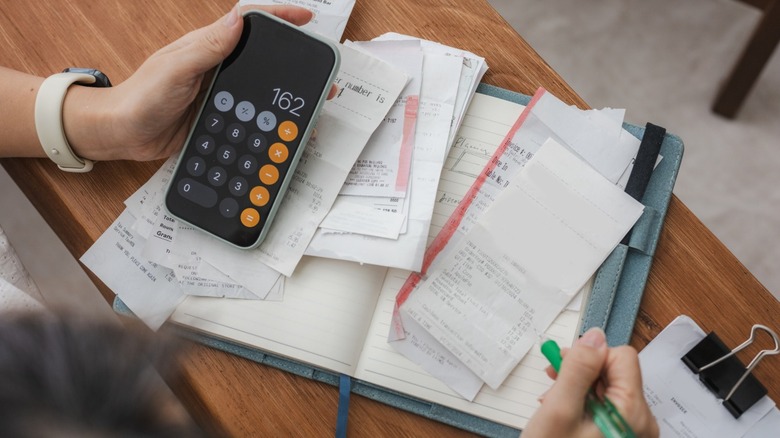 A person budgeting their household expenses with a calculator app on their phone and a stack of receipts.
