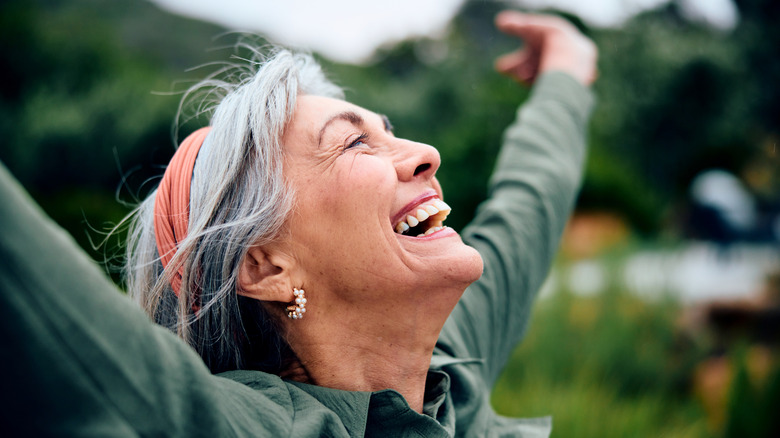 A closeup of a carefree retired woman's smiling face with her arms spread out toward the sky standing outside.