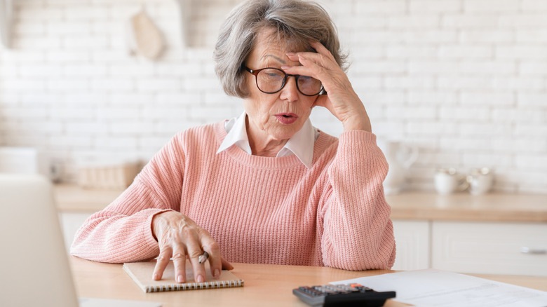 A retired woman sitting at a table, looking at a calculator with one hand on her head and the other on a notebook holding a pen.
