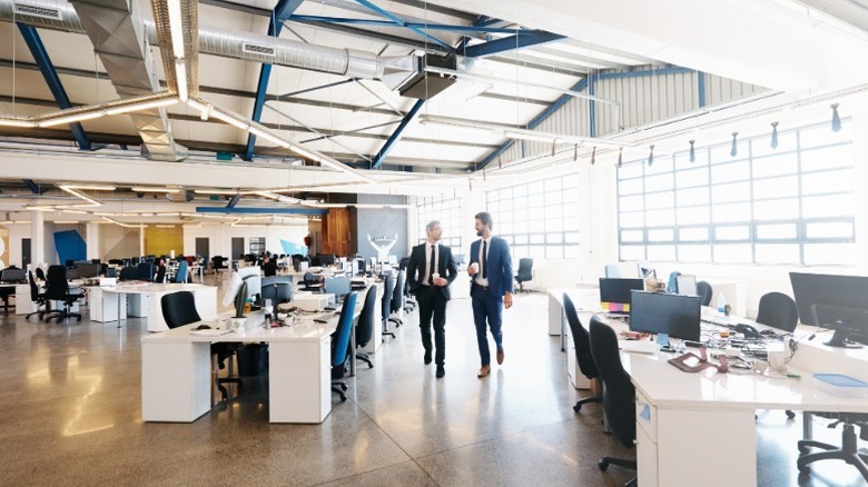 Two men in suits walking through an empty office that has multiple computer workstations on long white desks..