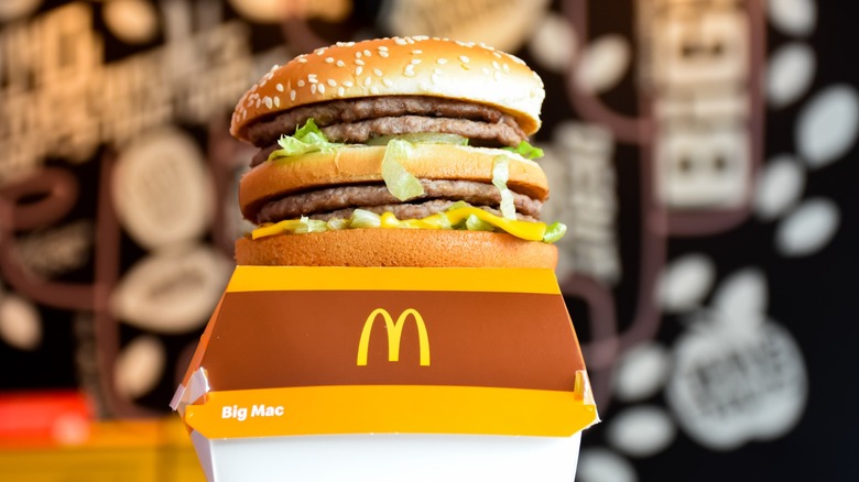 A McDonald's Big Mac in its open box, showing the stacked sesame bun, patties, lettuce, cheese, and special sauce against a blurred restaurant background