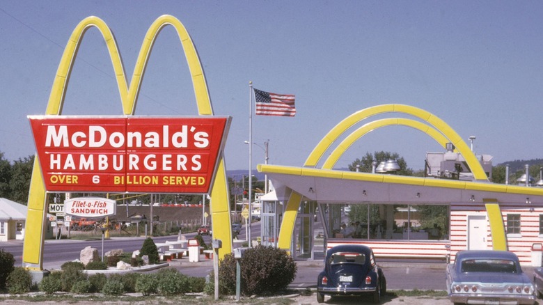 Vintage McDonald's restaurant with classic golden arches, a large red "Hamburgers—Over 6 Billion Served" sign, and two parked cars in front under a clear blue sky