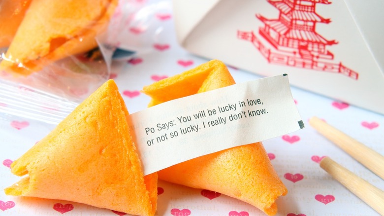 A typical fortune cookie with a slightly humorous or ominous fortune cracked open in front of a Chinese takeout box and other fortune cookie in a wrapper.