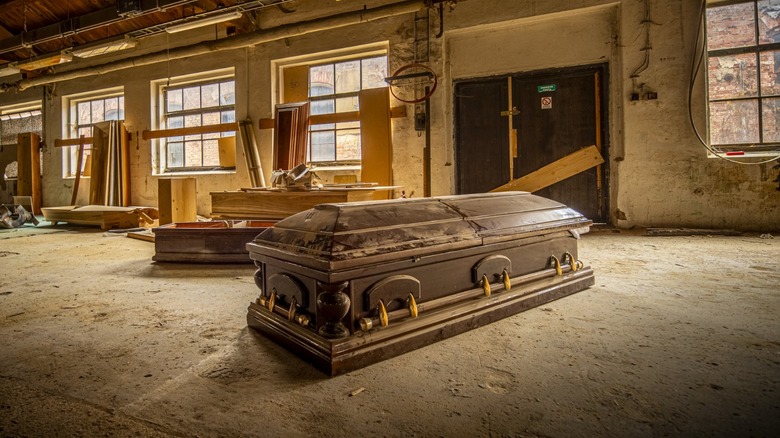Wooden coffin resting on a dusty workshop floor, surrounded by lumber and tools in a dimly lit industrial space