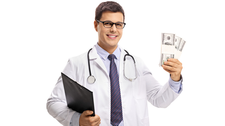 Smiling doctor with clipboard, stethoscope, and three stacks of $100 bills