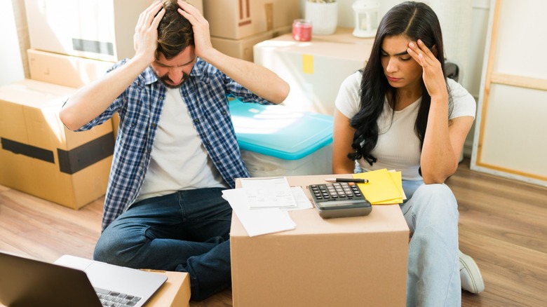 A stressed couple sits among moving boxes while reviewing bills with a calculator and laptop