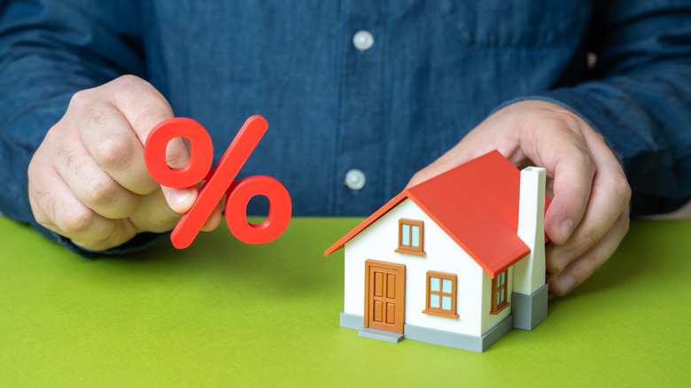 A close up of hands with one holding a red percentage symbol and the other holding a small white house figurine with a red roof.