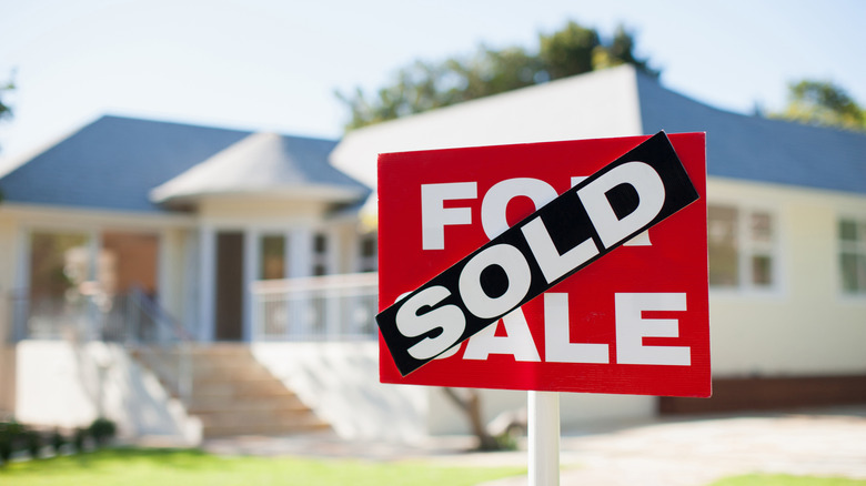A red for sale sign with a black sold sticker over it in front of a white house.