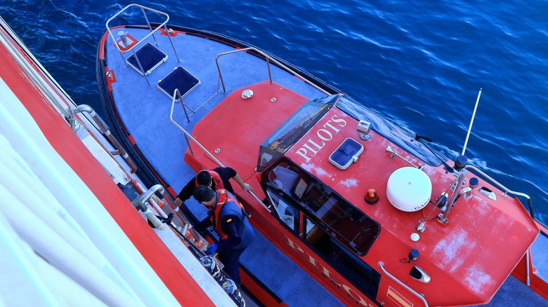 Two crew members prepare to transfer between a red pilot boat and a larger ship at sea.