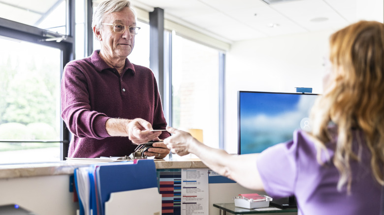 Old man gives card to woman sitting behind desk with folders and computer screen