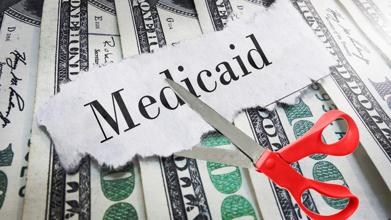 Scissors positioned to cut paper that says Medicaid on top of line of $100 bills