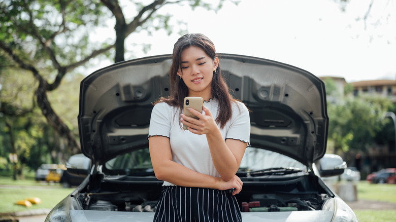 A woman waiting for roadside assistance in front of her car's open hood.