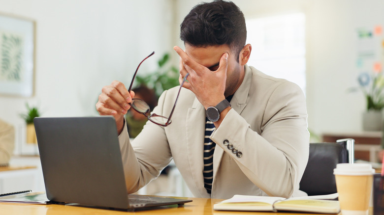 A man working on a laptop takes his glasses his off and rubs his eyes in frustration